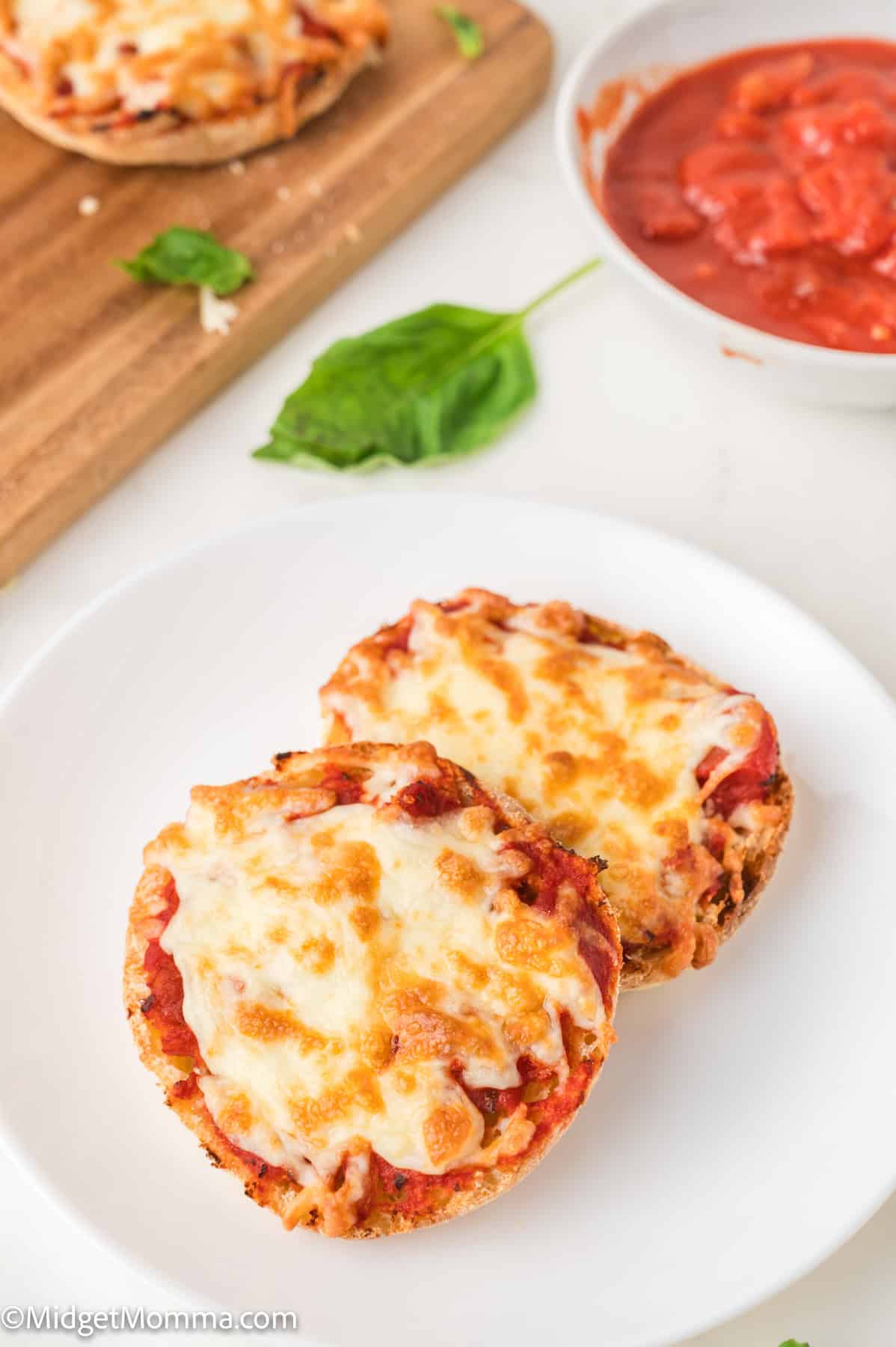 Two English muffin pizzas with melted cheese on a white plate, with a bowl of tomato sauce and basil leaves nearby.