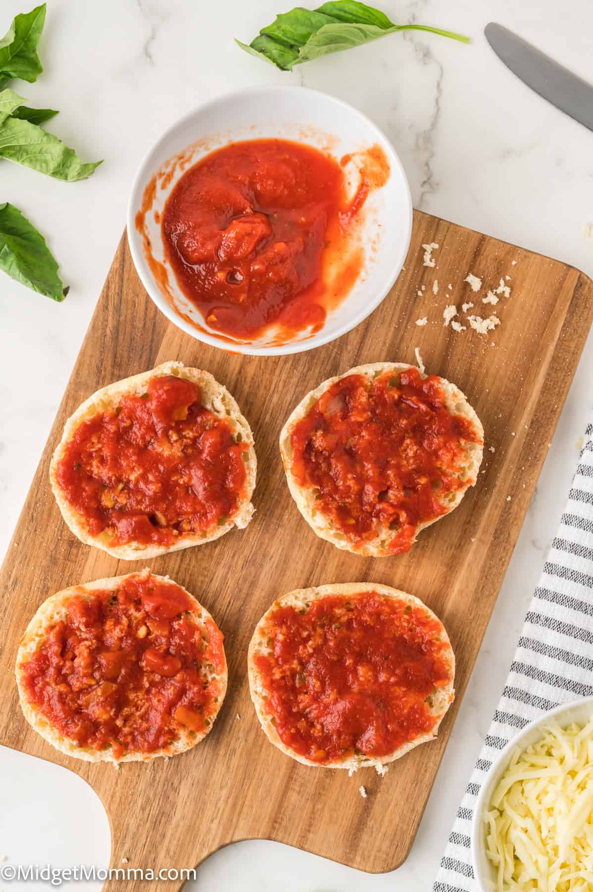 Four English muffin halves topped with tomato sauce on a wooden board, next to a bowl of sauce and fresh basil leaves.