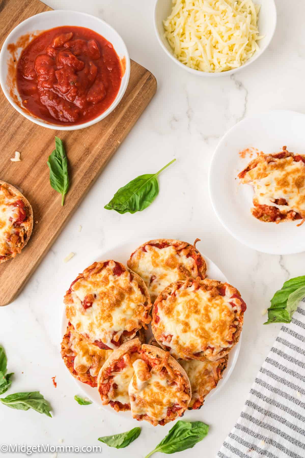 Mini pizza made with english muffins with melted cheese on a plate, accompanied by a bowl of tomato sauce, a bowl of shredded cheese, and fresh basil leaves on a white surface.