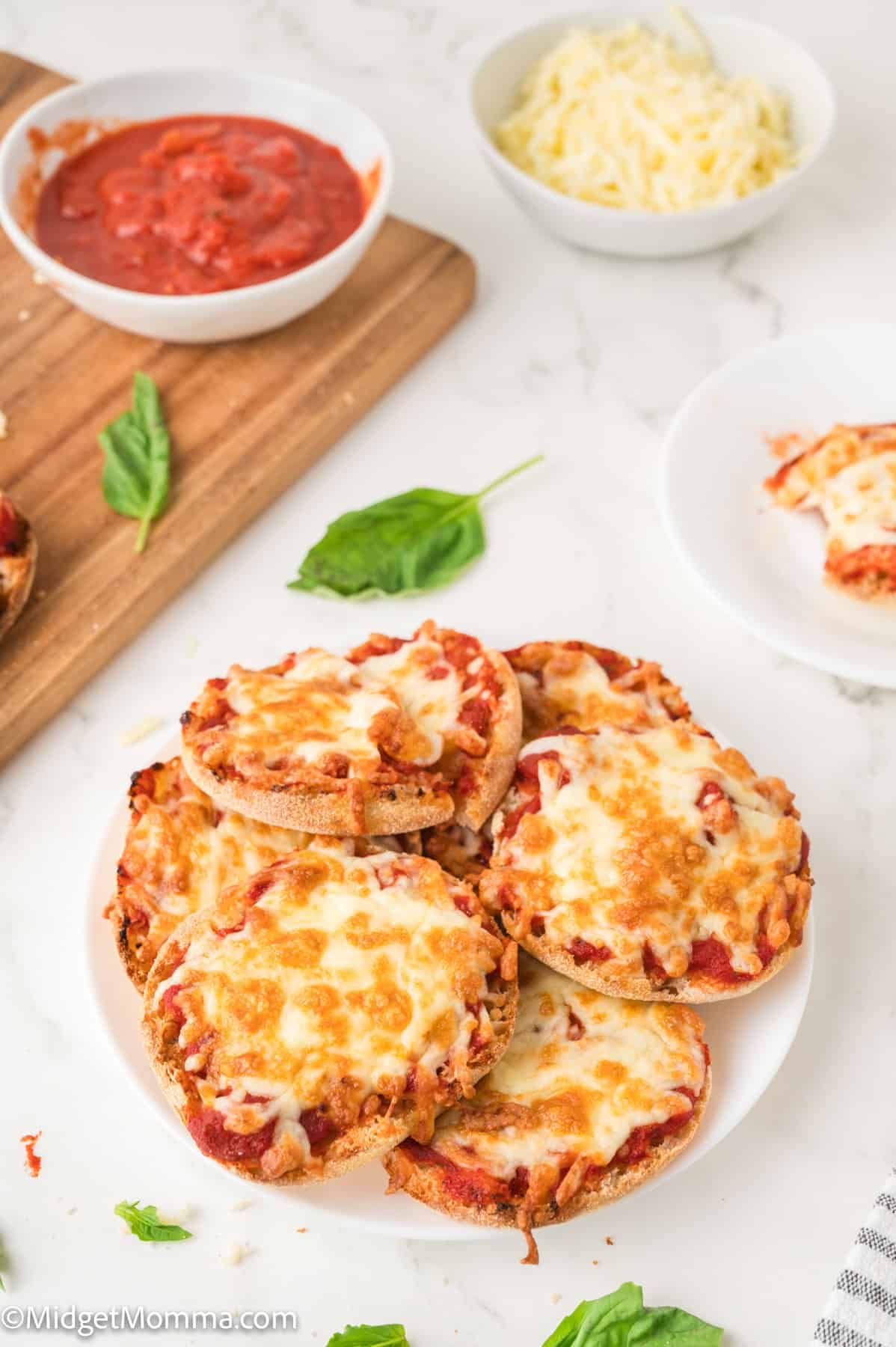 Plate of homemade English muffin pizzas topped with melted cheese, on a marble surface. Nearby are a bowl of tomato sauce and shredded cheese, with basil leaves scattered around.