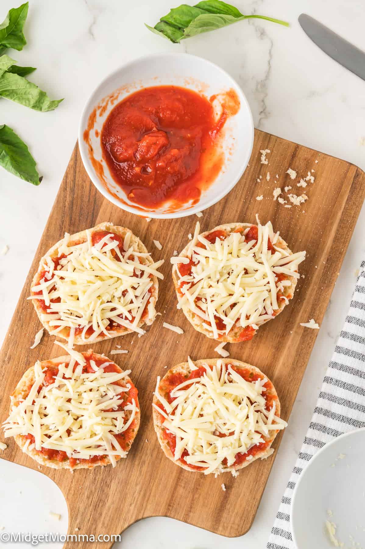 Four English muffin halves topped with tomato sauce and shredded cheese sit on a wooden board. A bowl of tomato sauce and basil leaves are nearby.