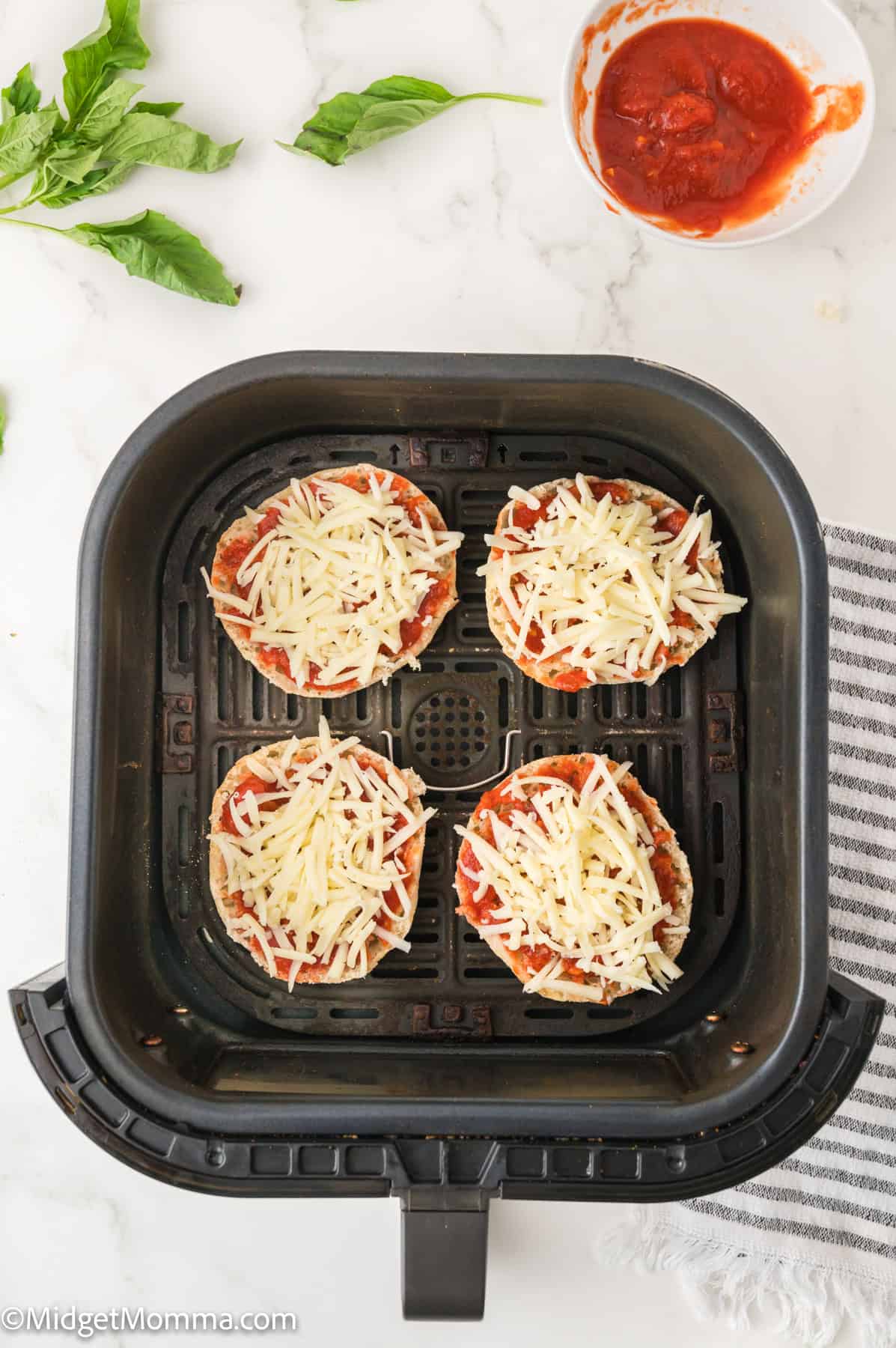 Air fryer basket with four uncooked eggplant slices topped with tomato sauce and shredded cheese. Bowl of sauce and basil leaves nearby.