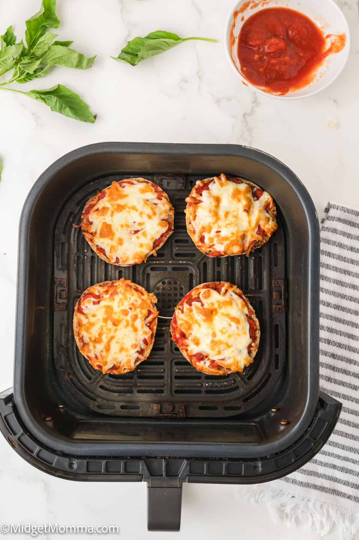 Four mini pizzas with melted cheese in an air fryer basket, with a bowl of tomato sauce and fresh basil leaves nearby.