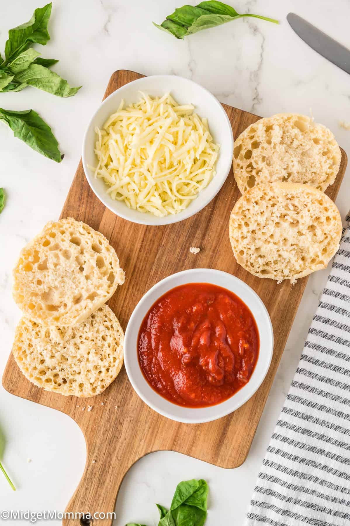 Wooden board with split English muffins, a bowl of shredded cheese, and a bowl of tomato sauce. Basil leaves and a striped cloth napkin are nearby.