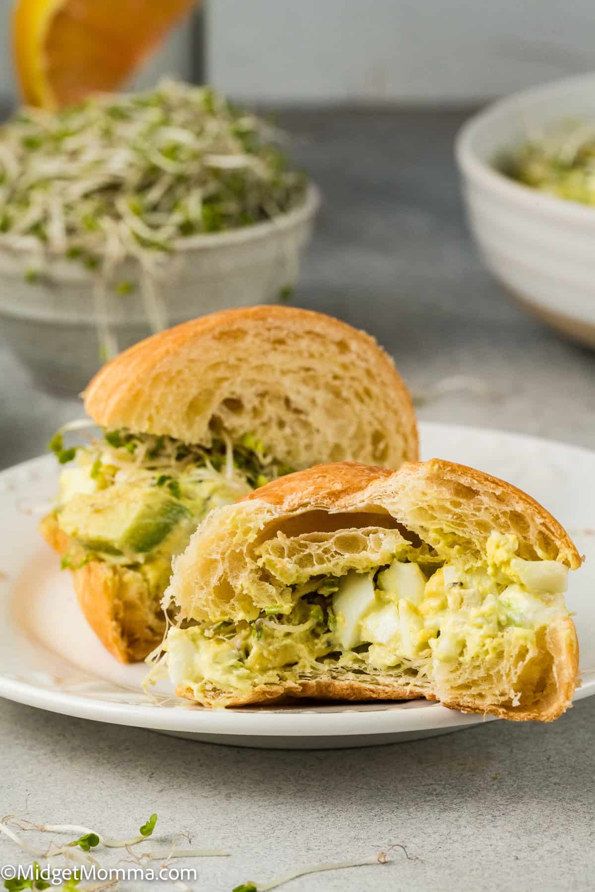 Cross-section of a sandwich with avocado, egg salad, and sprouts on a white plate.