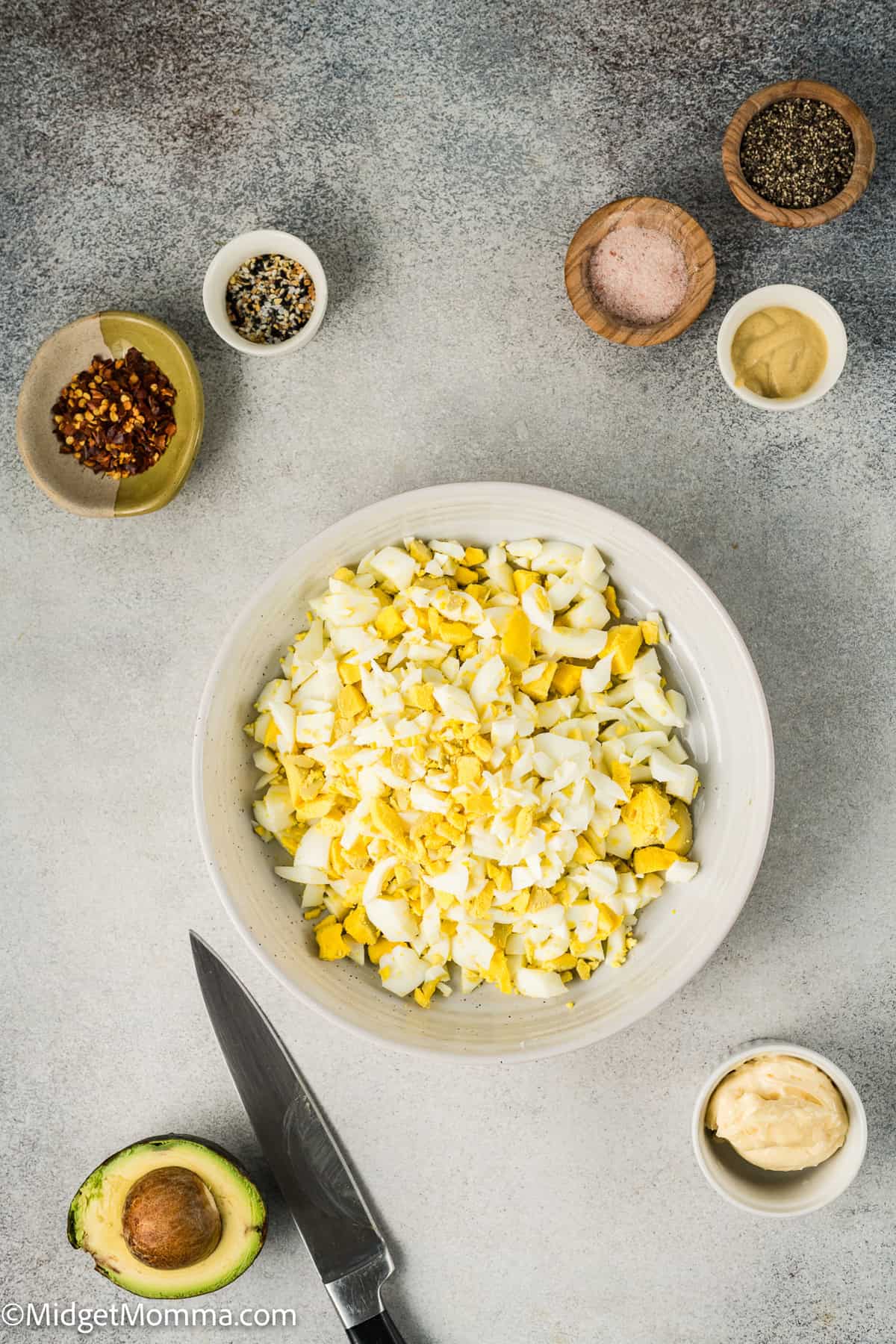 Chopped boiled eggs in a bowl surrounded by seasonings, avocado, a knife, and a dollop of mayonnaise on a gray surface.