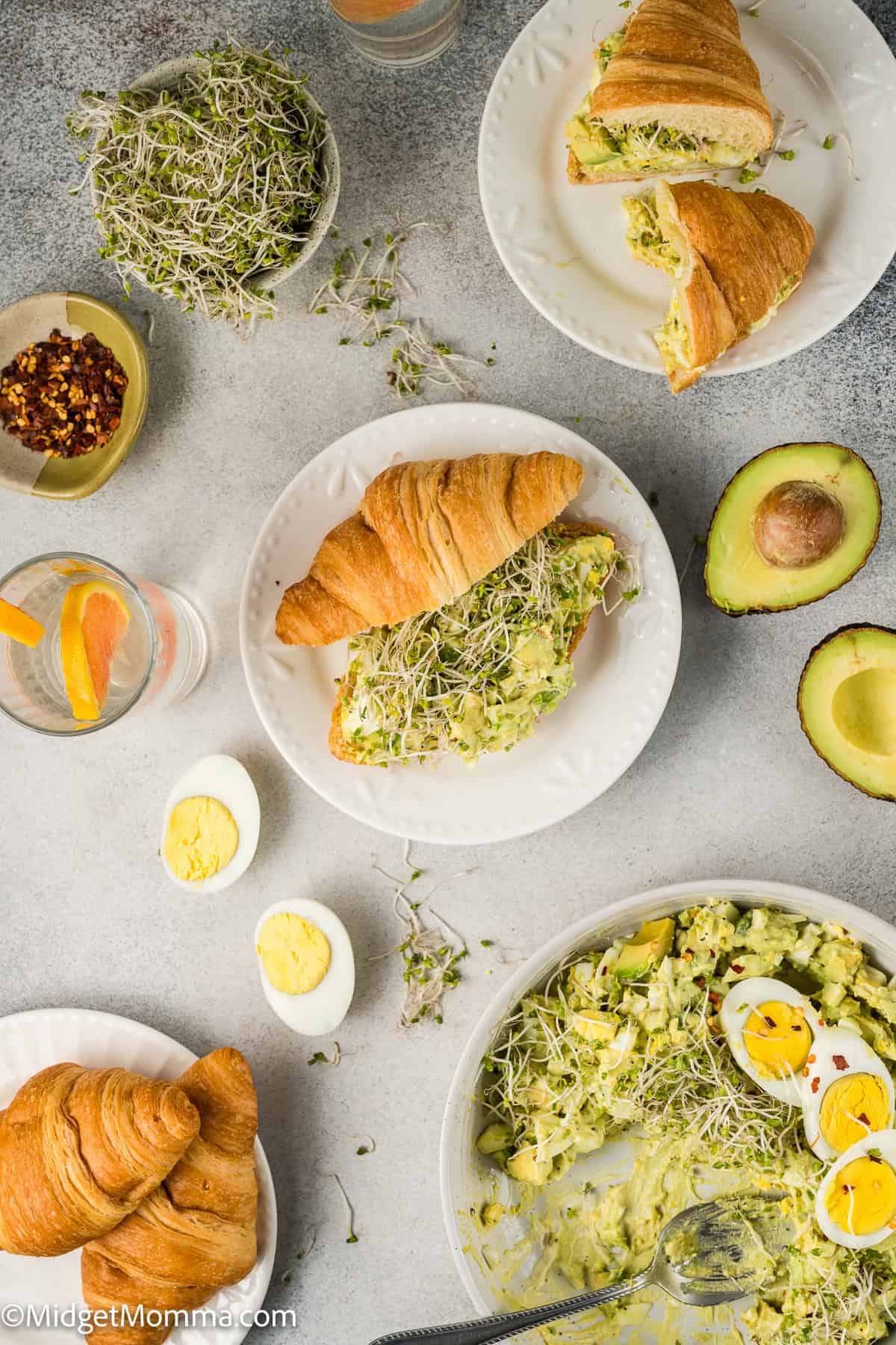 Croissants with avocado egg salad and sprouts on plates, surrounded by halved avocado, hard-boiled eggs, red pepper flakes, and drinks with orange slices.