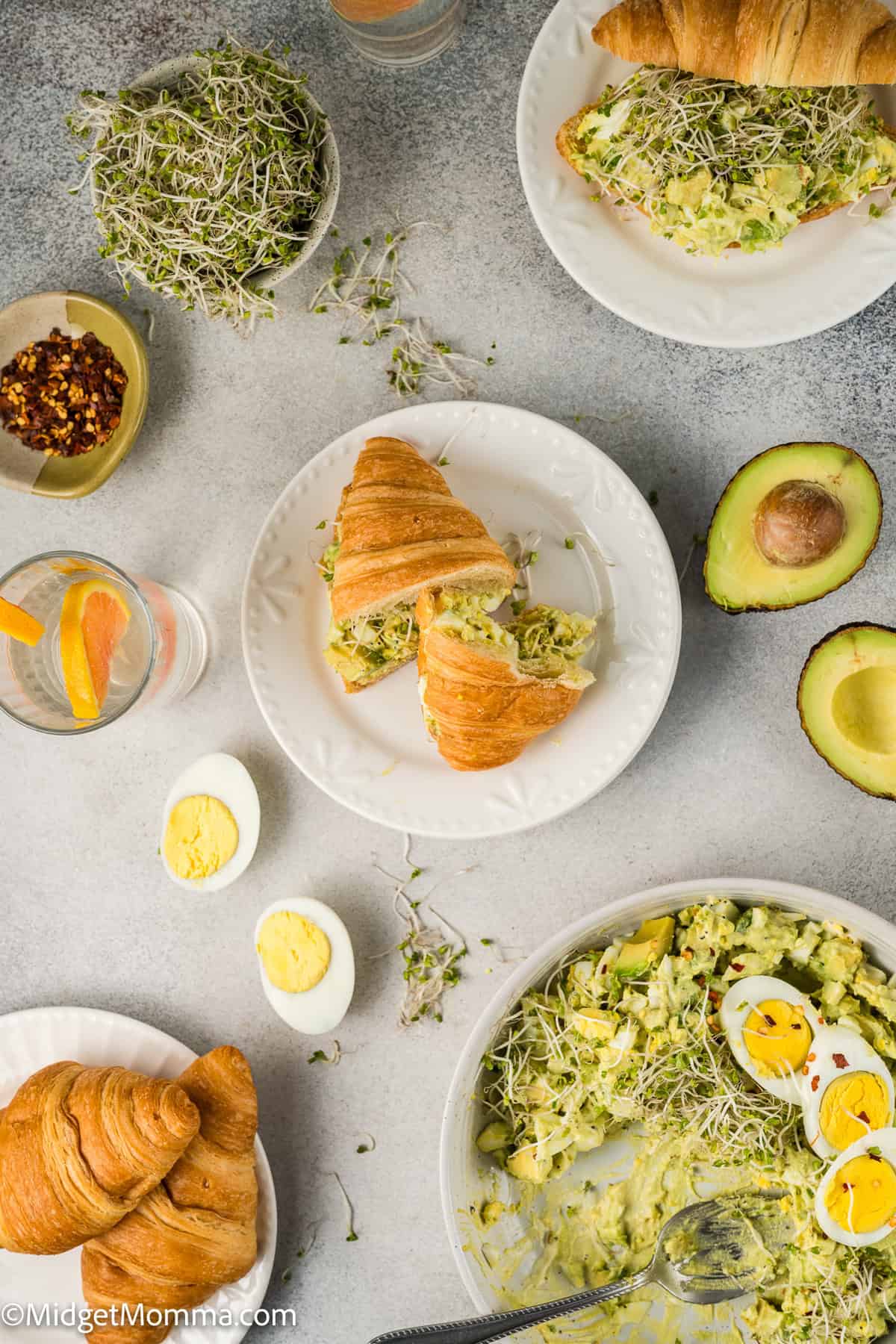 Croissants filled with avocado egg salad on plates, surrounded by ingredients like alfalfa sprouts, hard-boiled eggs, avocado halves, and glasses of water with lemon.