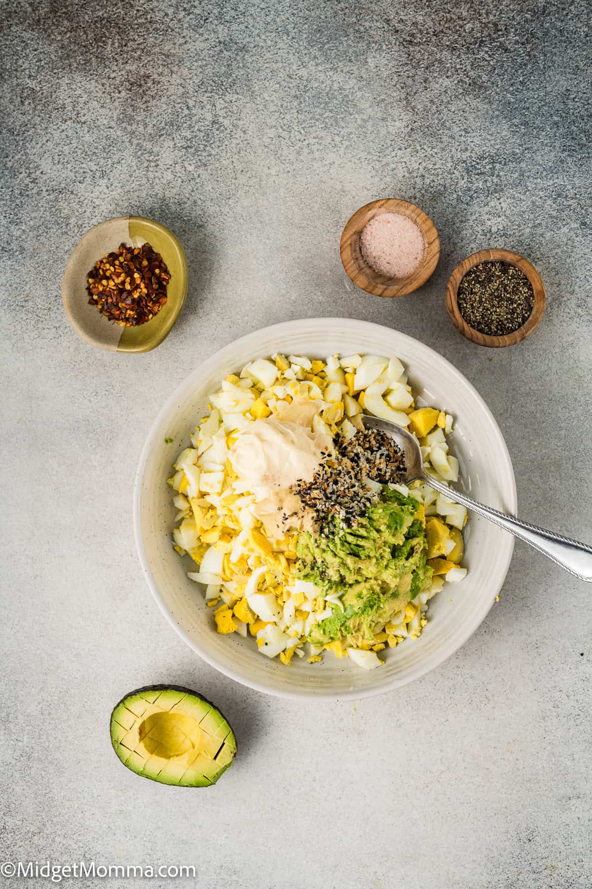 Bowl of chopped eggs, shredded avocado, and seasoning on a table with a spoon. Surrounding bowls contain red pepper flakes, pink salt, and black pepper. Half an avocado is nearby.