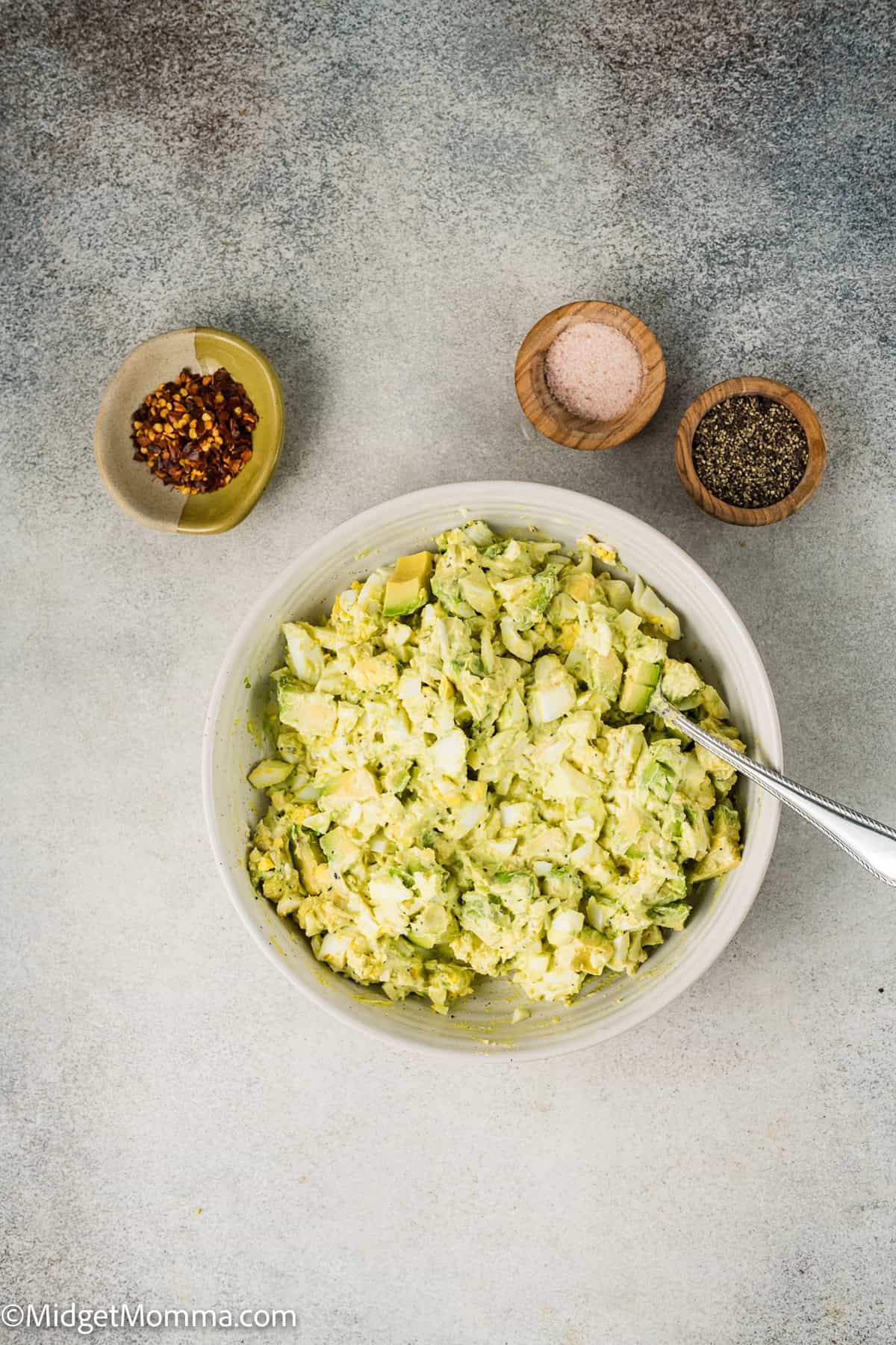 A bowl of egg salad with a spoon on a textured surface, surrounded by small bowls containing red pepper flakes, pink salt, and black pepper.