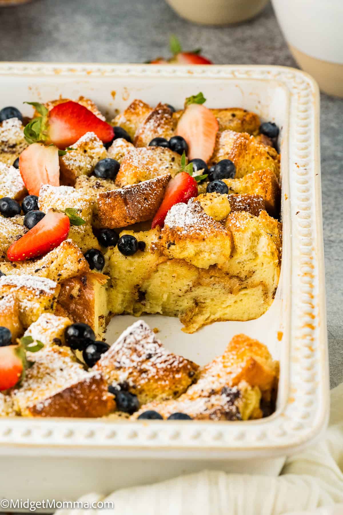 Close-up of French toast topped with strawberries, blueberries, and powdered sugar, with syrup being drizzled on top. A fork is placed beside the toast on a white plate.