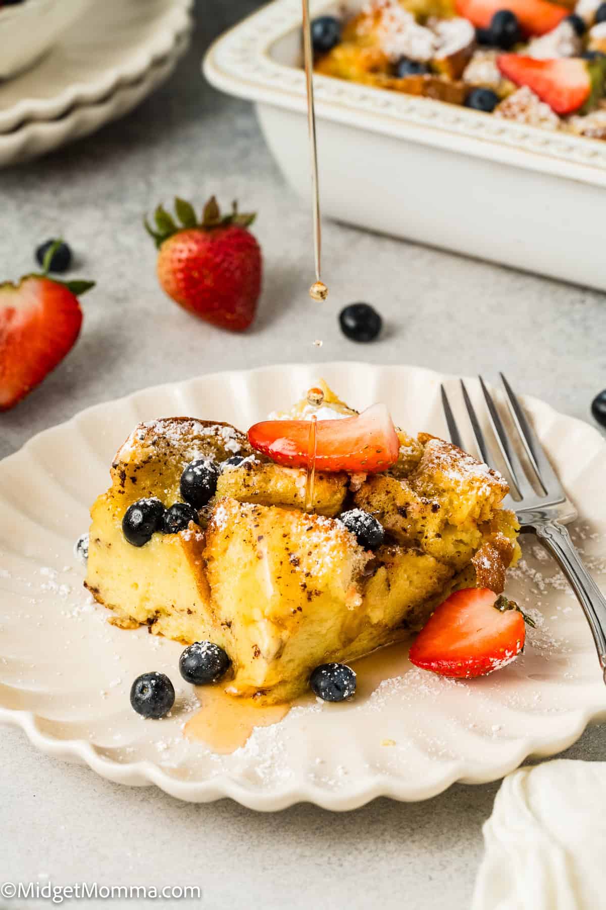 Close-up of French toast topped with strawberries, blueberries, and powdered sugar, with syrup being drizzled on top. A fork is placed beside the toast on a white plate.