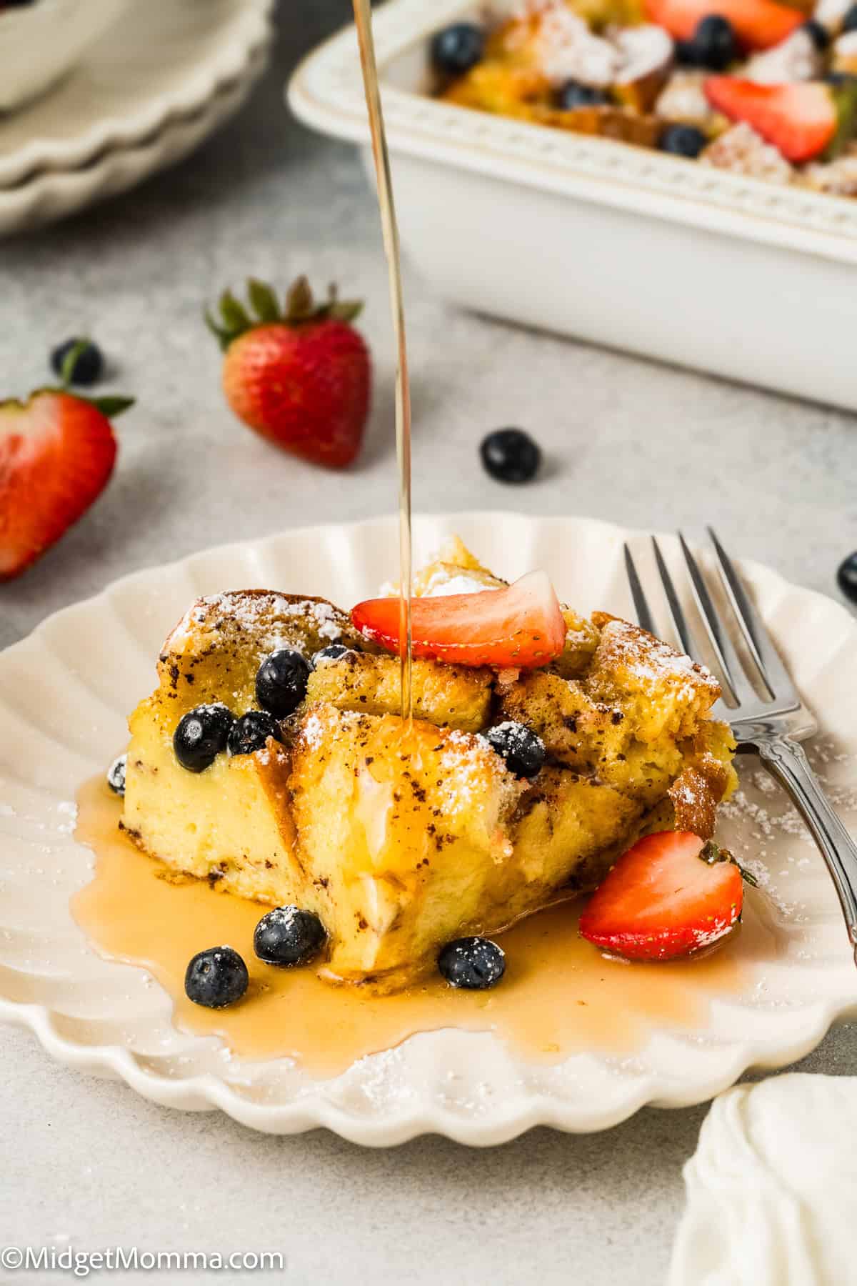 A slice of baked French toast casserole topped with strawberries and blueberries, with syrup being poured over it, on a white plate next to a fork.