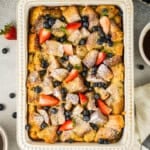 Baked French toast casserole topped with powdered sugar, strawberries, and blueberries in a rectangular dish, surrounded by fresh fruit and a cup of coffee.