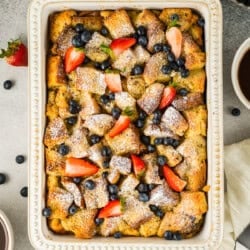 Baked French toast casserole topped with powdered sugar, strawberries, and blueberries in a rectangular dish, surrounded by fresh fruit and a cup of coffee.