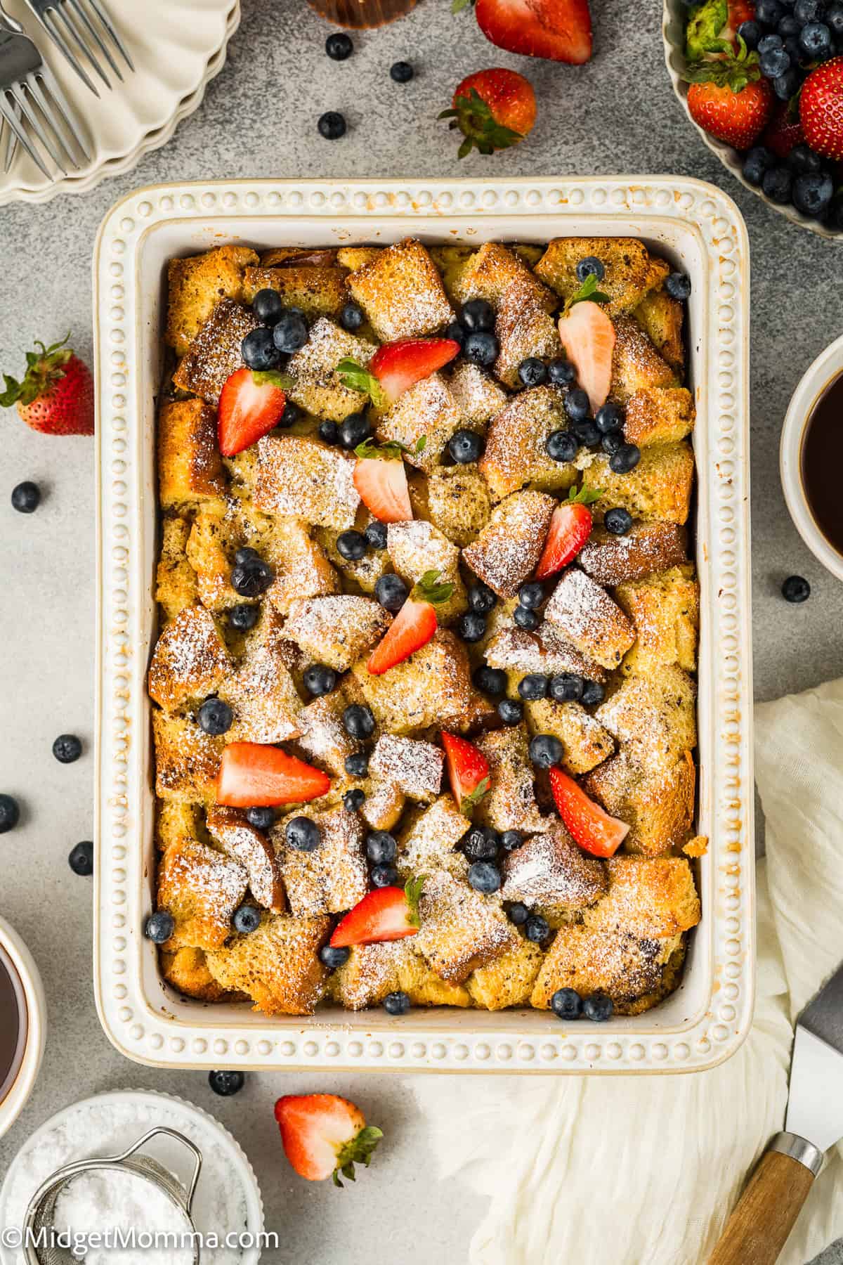 Baked French toast casserole topped with powdered sugar, strawberries, and blueberries in a rectangular dish, surrounded by fresh fruit and a cup of coffee.