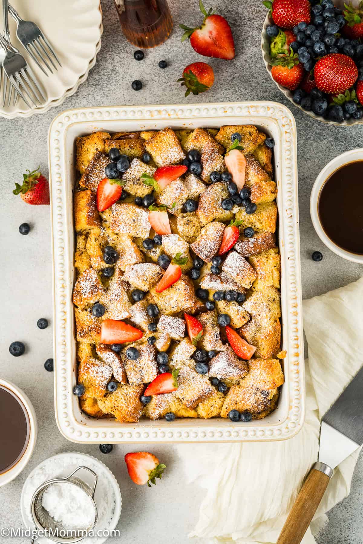 A rectangular dish with baked French toast topped with powdered sugar, strawberries, and blueberries. Surrounding items include cups of coffee, a knife, whipped cream, and a bowl of berries.