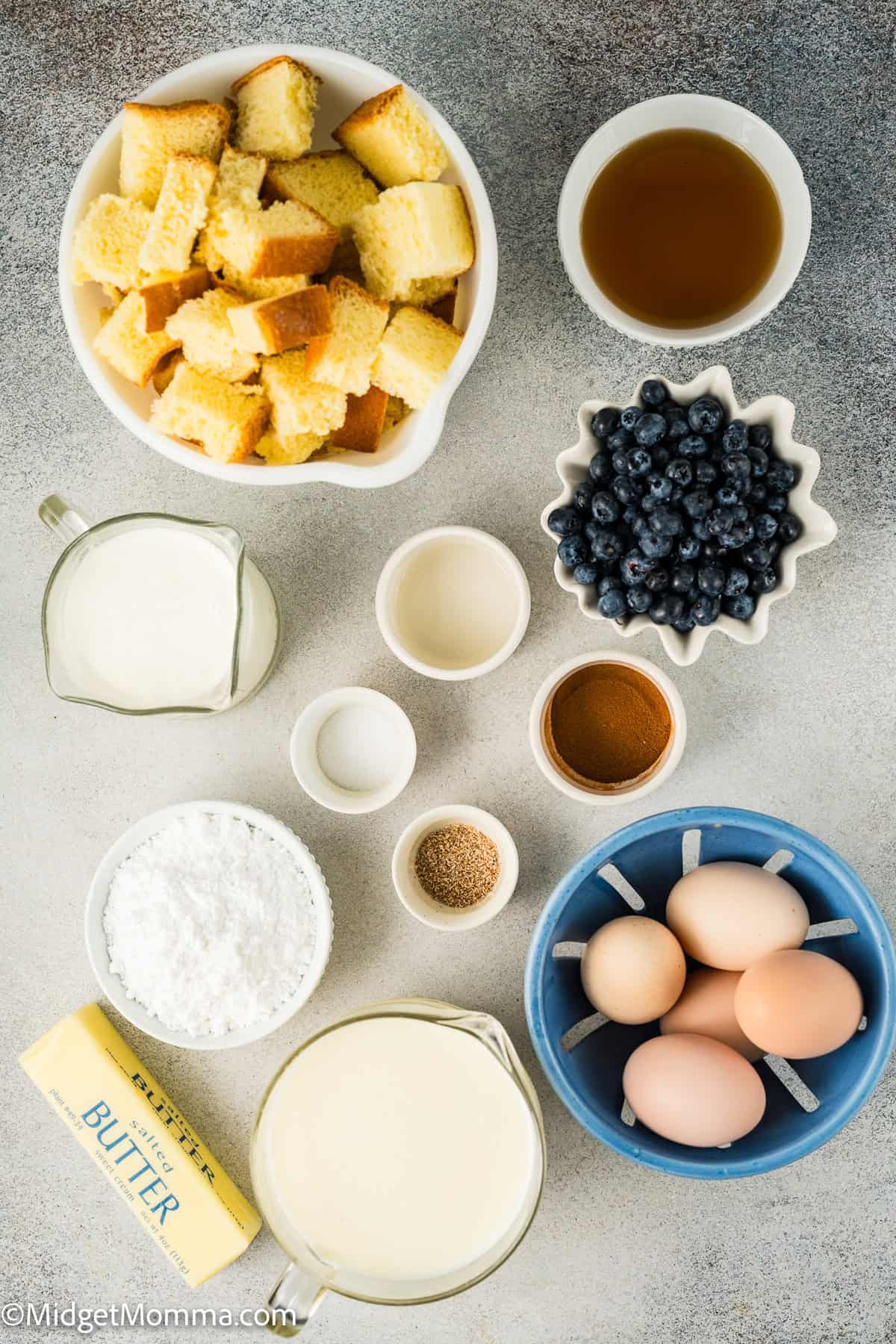 brioche french toast casserole ingredients. Ingredients for a recipe are arranged on a table: cubed bread, blueberries, eggs, butter, cream, milk, sugar, cinnamon, vanilla extract, and nutmeg.