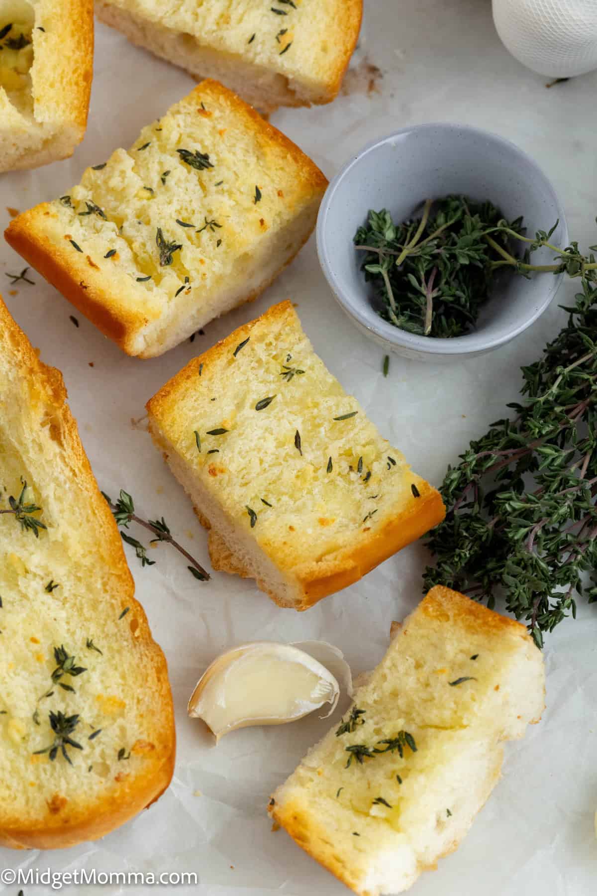 Slices of garlic bread with herbs, a sprig of fresh thyme, a garlic clove, and a small bowl with loose herbs on a white surface.