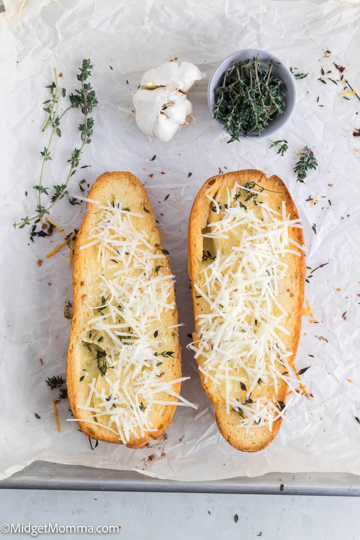 Two slices of garlic bread with grated cheese on parchment paper, surrounded by fresh thyme sprigs and a bulb of garlic.