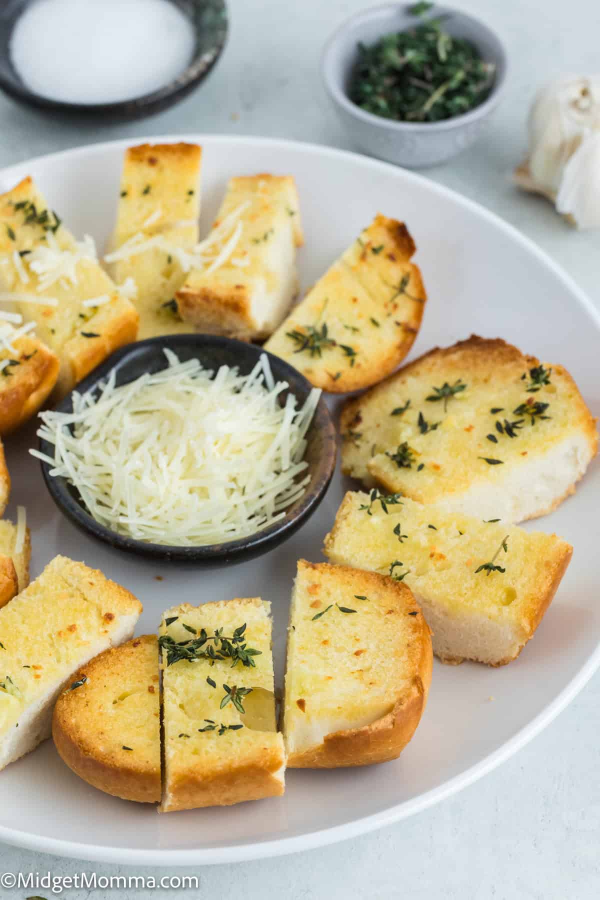 A plate of sliced garlic bread with herbs, arranged around a small bowl of shredded cheese, next to salt and chopped herbs.