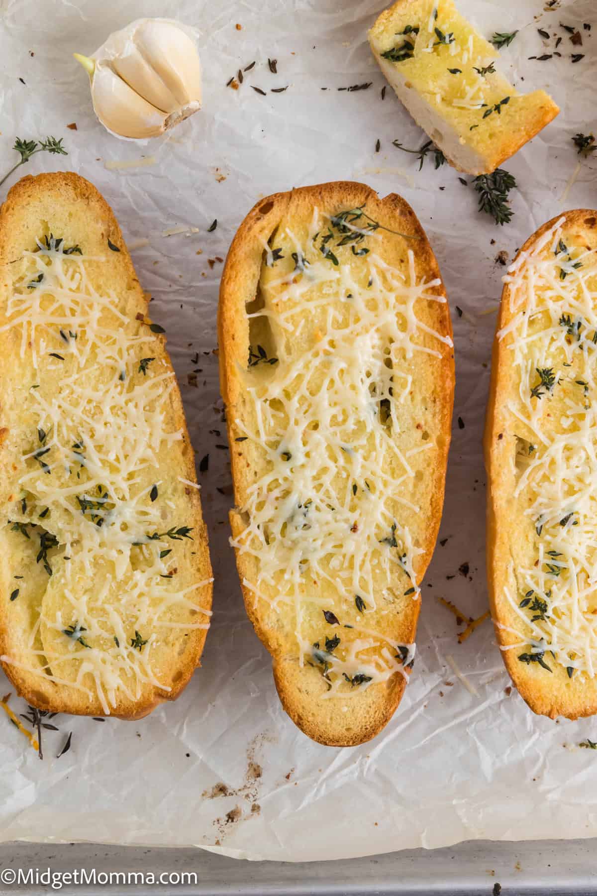 Three slices of garlic bread topped with grated cheese and herbs, placed on parchment paper, with a garlic bulb nearby.