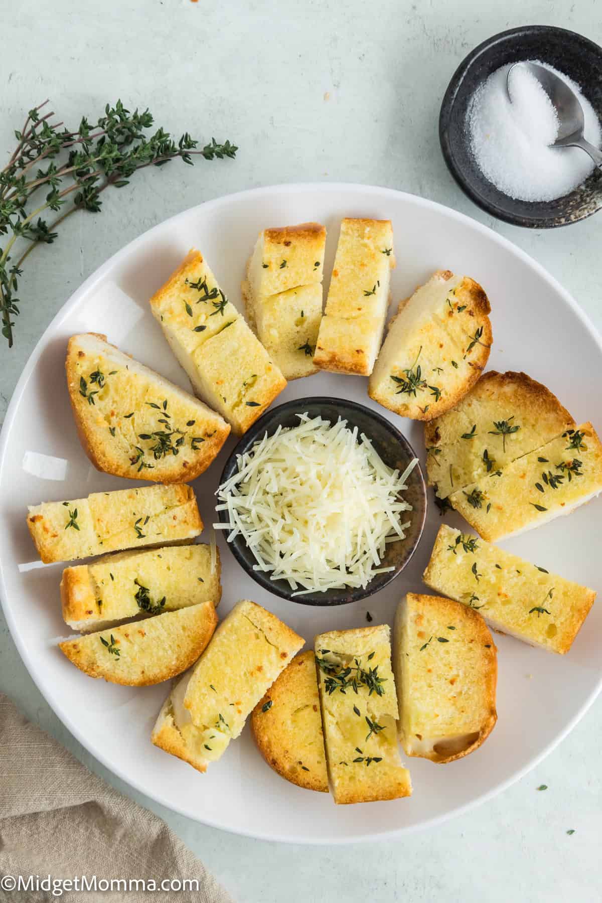 Slices of garlic bread with herbs arranged in a circle around a bowl of grated cheese on a white plate. Salt in a small dish nearby.