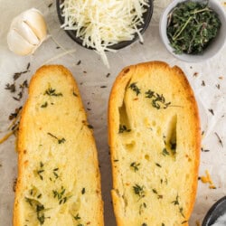 Two slices of garlic bread with herbs, alongside bowls of grated cheese, herbs, and a garlic bulb on parchment paper.