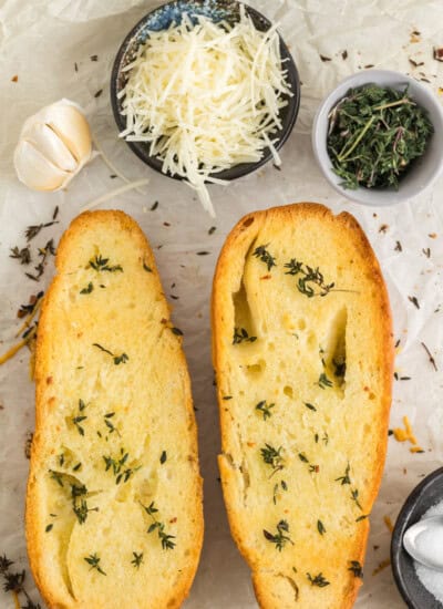 Two slices of garlic bread with herbs, alongside bowls of grated cheese, herbs, and a garlic bulb on parchment paper.