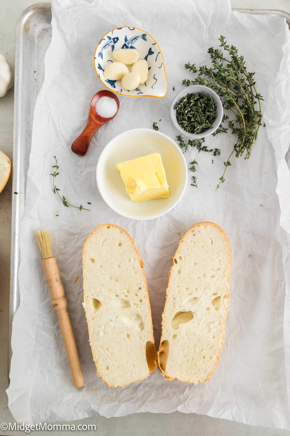 Sliced bread, butter, garlic cloves, a wooden brush, and herbs on parchment paper.