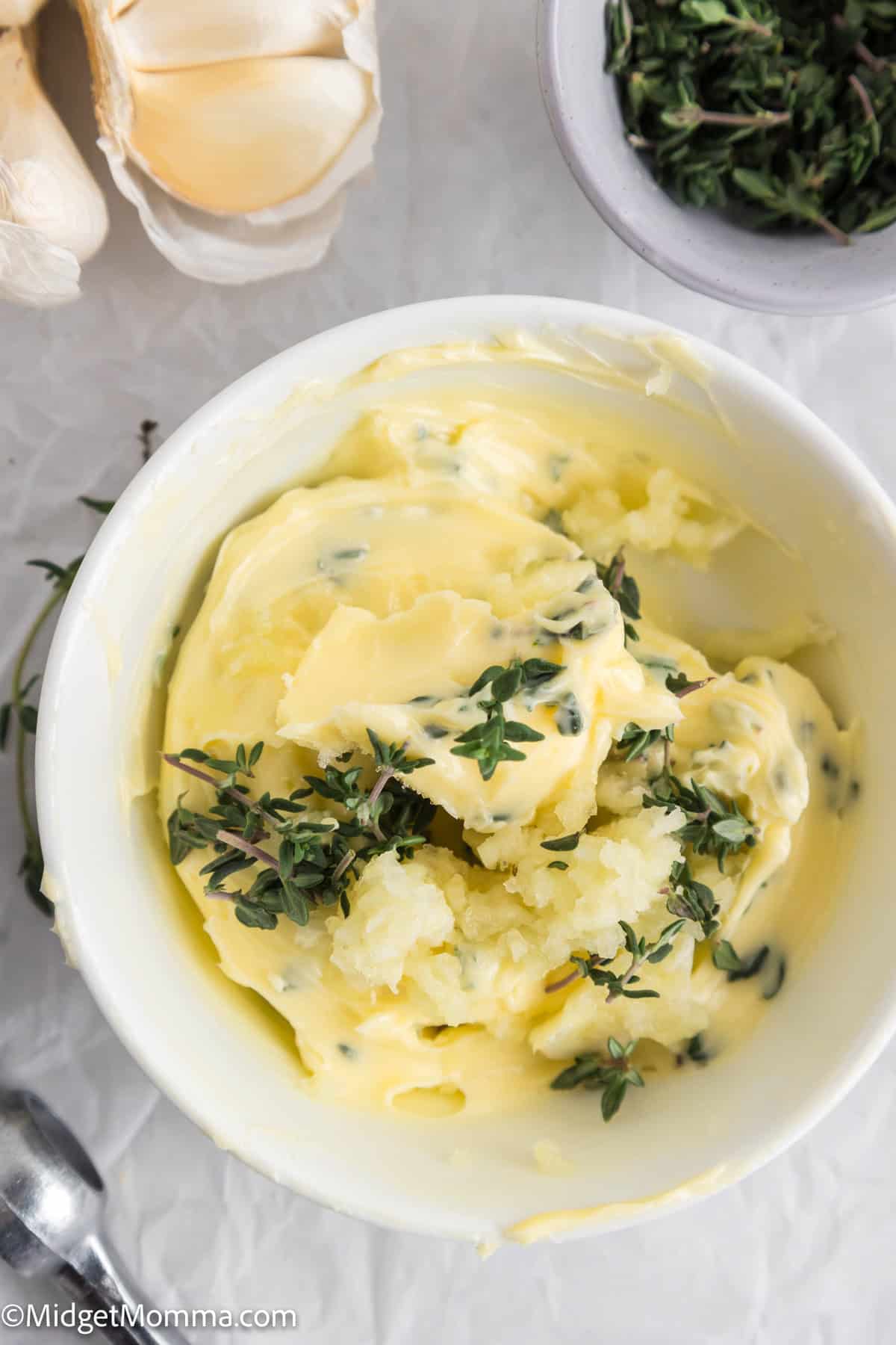 A bowl of herb butter with fresh thyme on top, next to garlic cloves and a small bowl of chopped herbs.