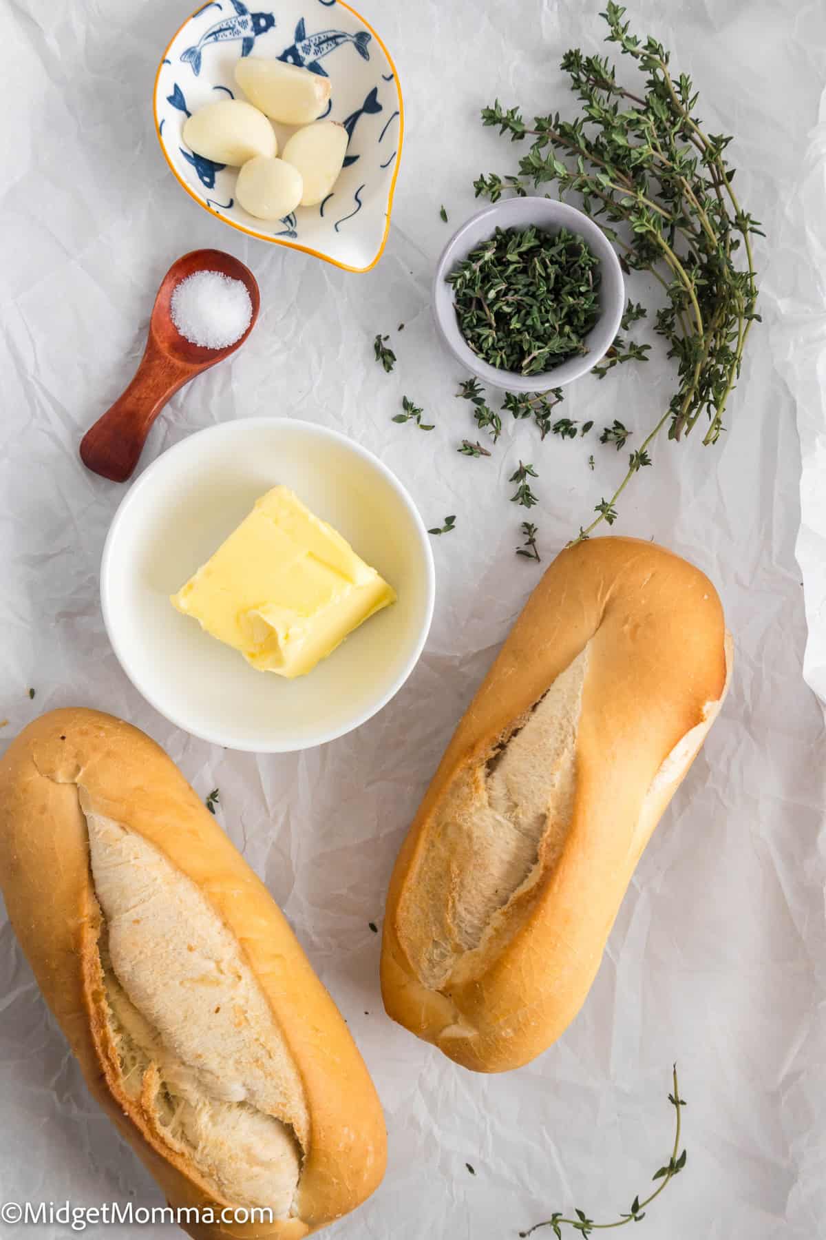 Homemade Garlic Bread with Thyme Ingredients. Two baguettes, butter, garlic cloves, a small bowl of herbs, salt, and fresh thyme sprigs arranged on crumpled parchment paper. 