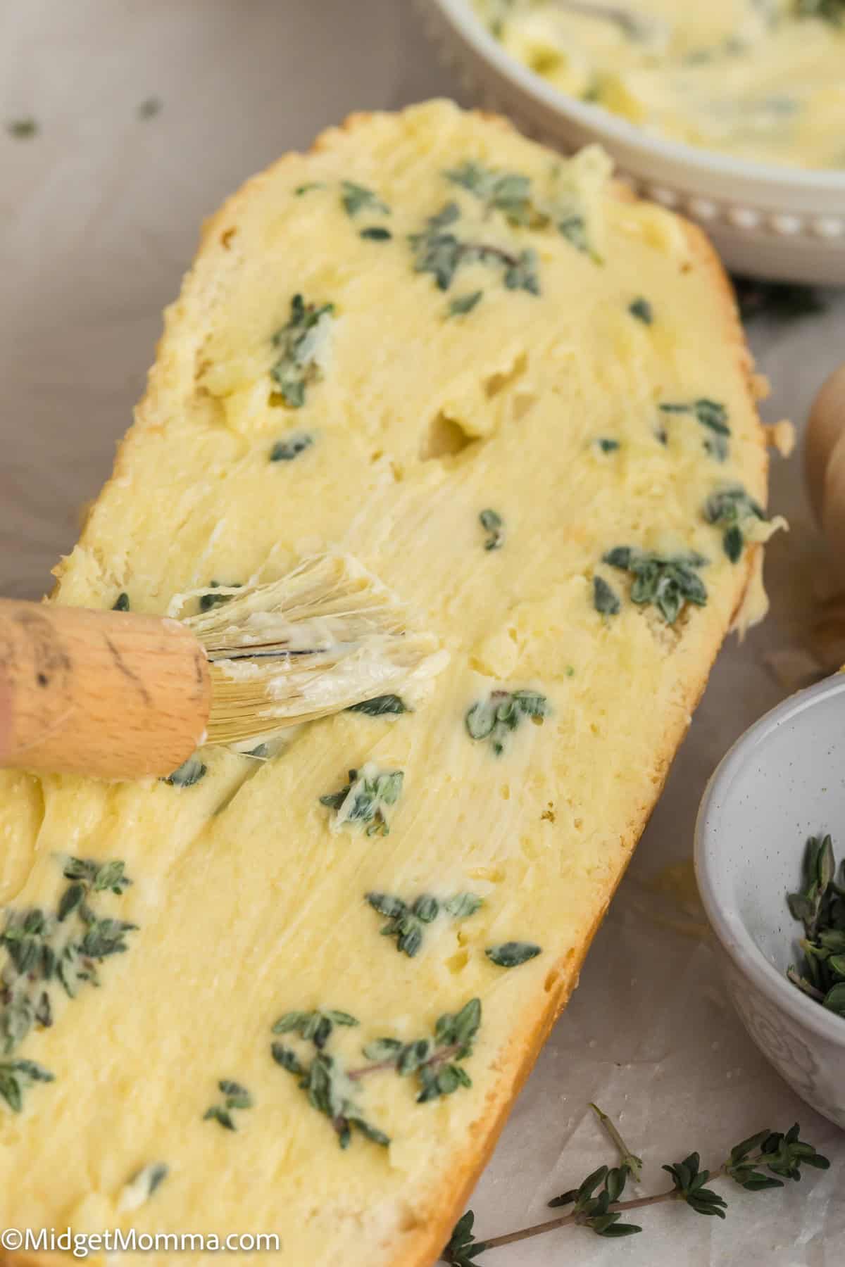 A slice of bread is being brushed with a buttery spread and topped with herbs.