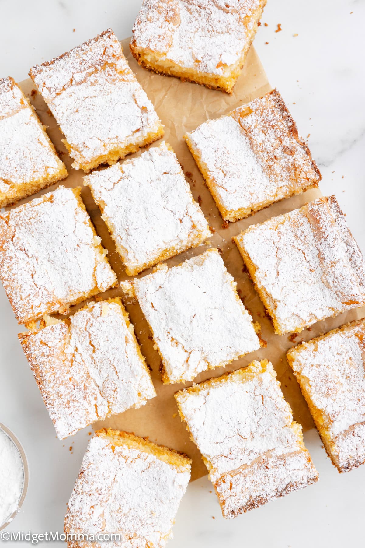 Twelve square slices of cake topped with powdered sugar are arranged on a sheet of parchment paper on a marble surface.