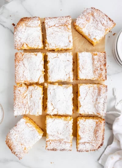 Twelve powdered sugar-topped cake squares arranged on parchment paper on a marble surface.