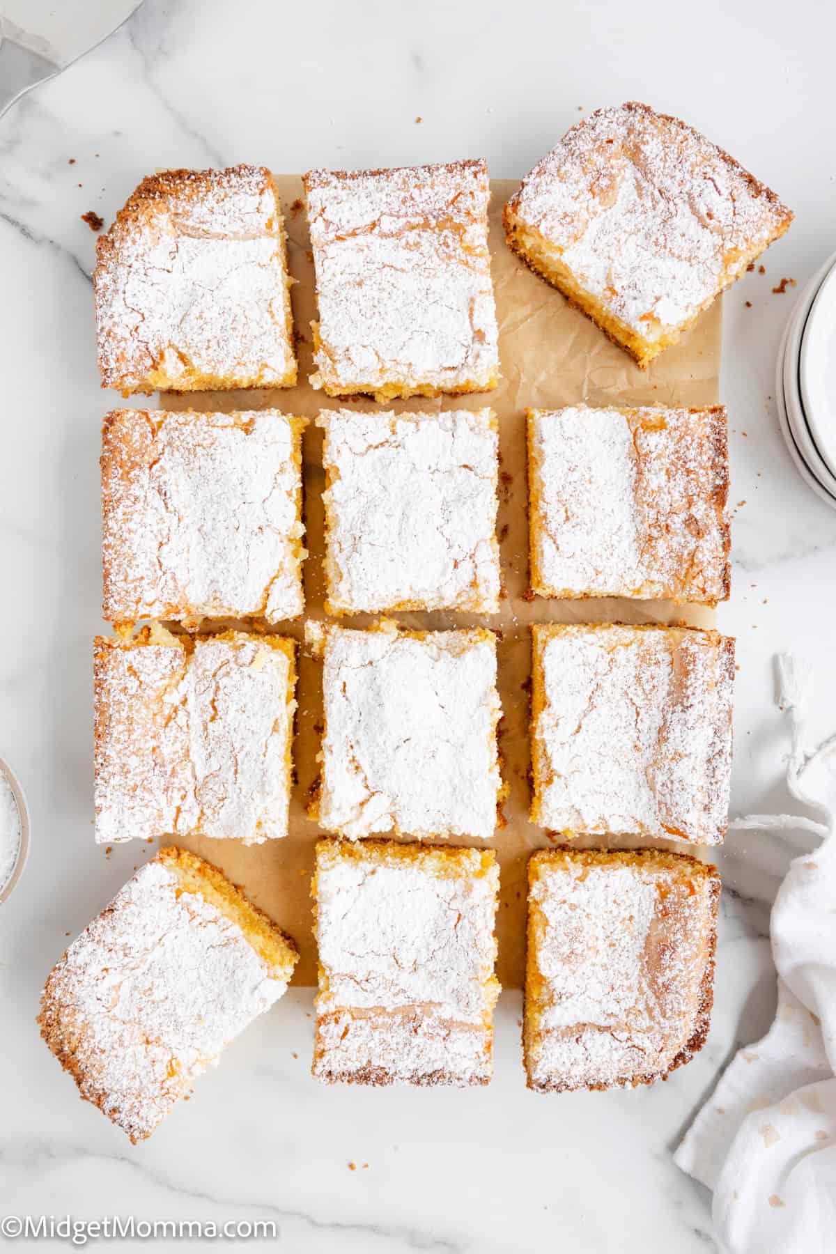 Twelve powdered sugar-topped cake squares arranged on parchment paper on a marble surface.