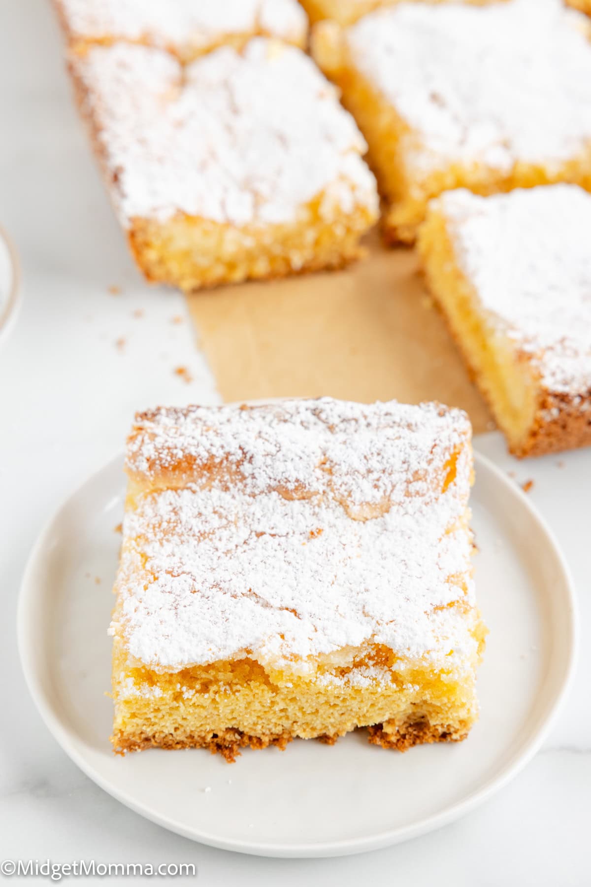 A piece of powdered sugar-covered cake on a plate, with more cake pieces in the background on parchment paper.