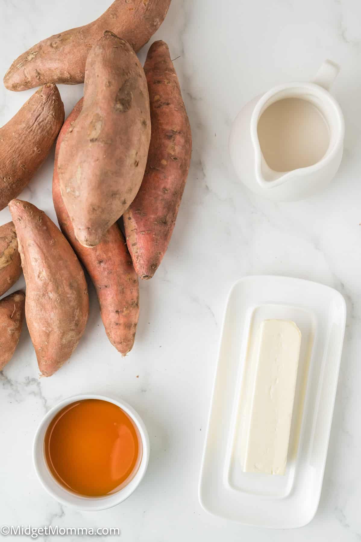 Sweet potatoes, a stick of butter on a plate, a pitcher of milk, and a bowl of orange liquid on a white surface.