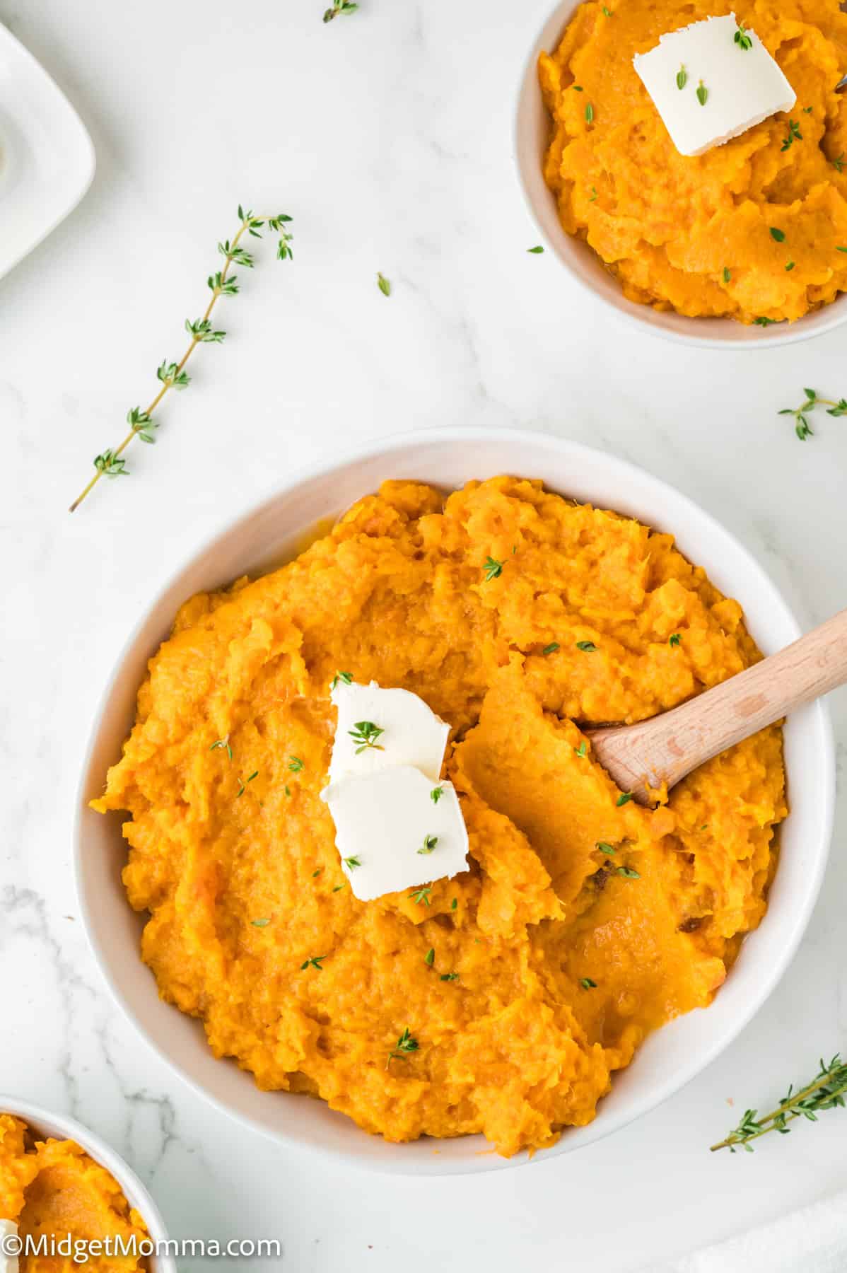 Mashed sweet potatoes in a bowl with two butter pats and a wooden spoon, garnished with fresh thyme.