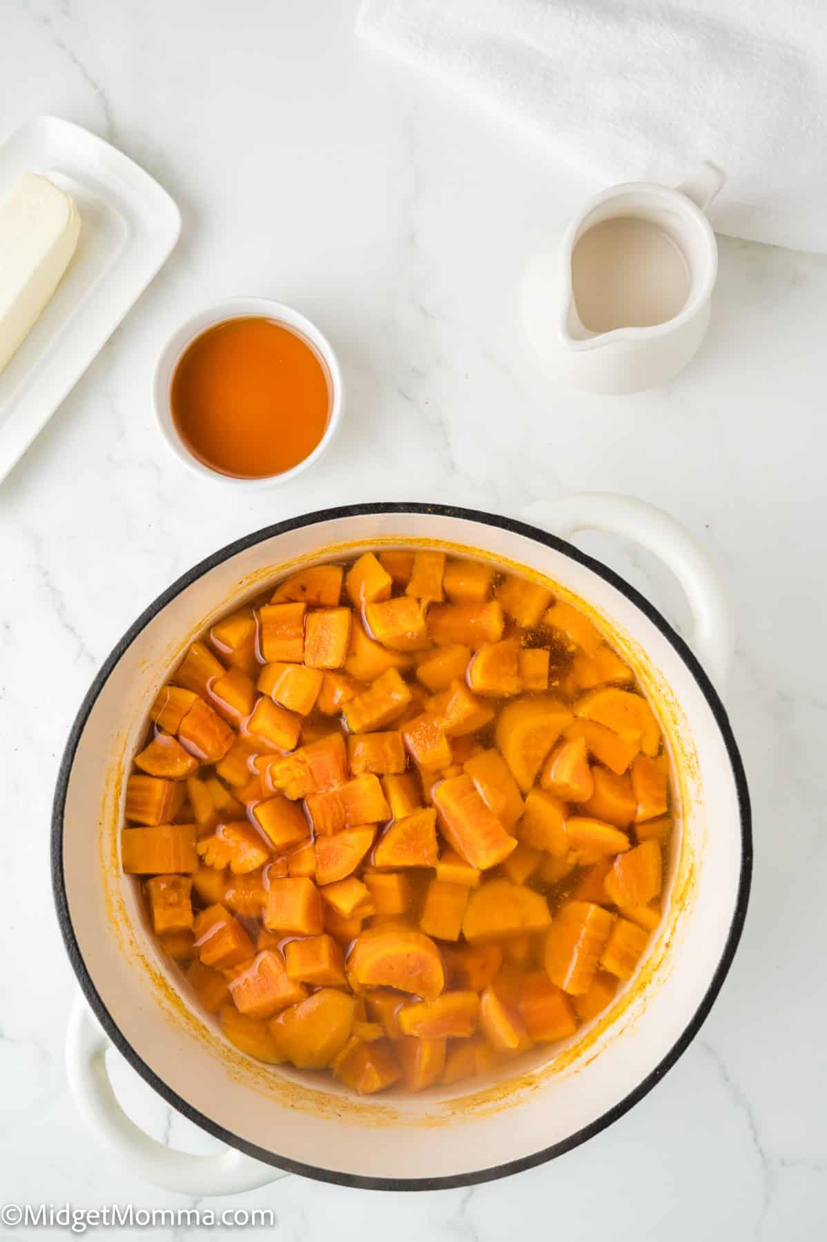 Top view of a pot with sliced yams in liquid, surrounded by small bowls of butter, sauce, and cream on a white surface.