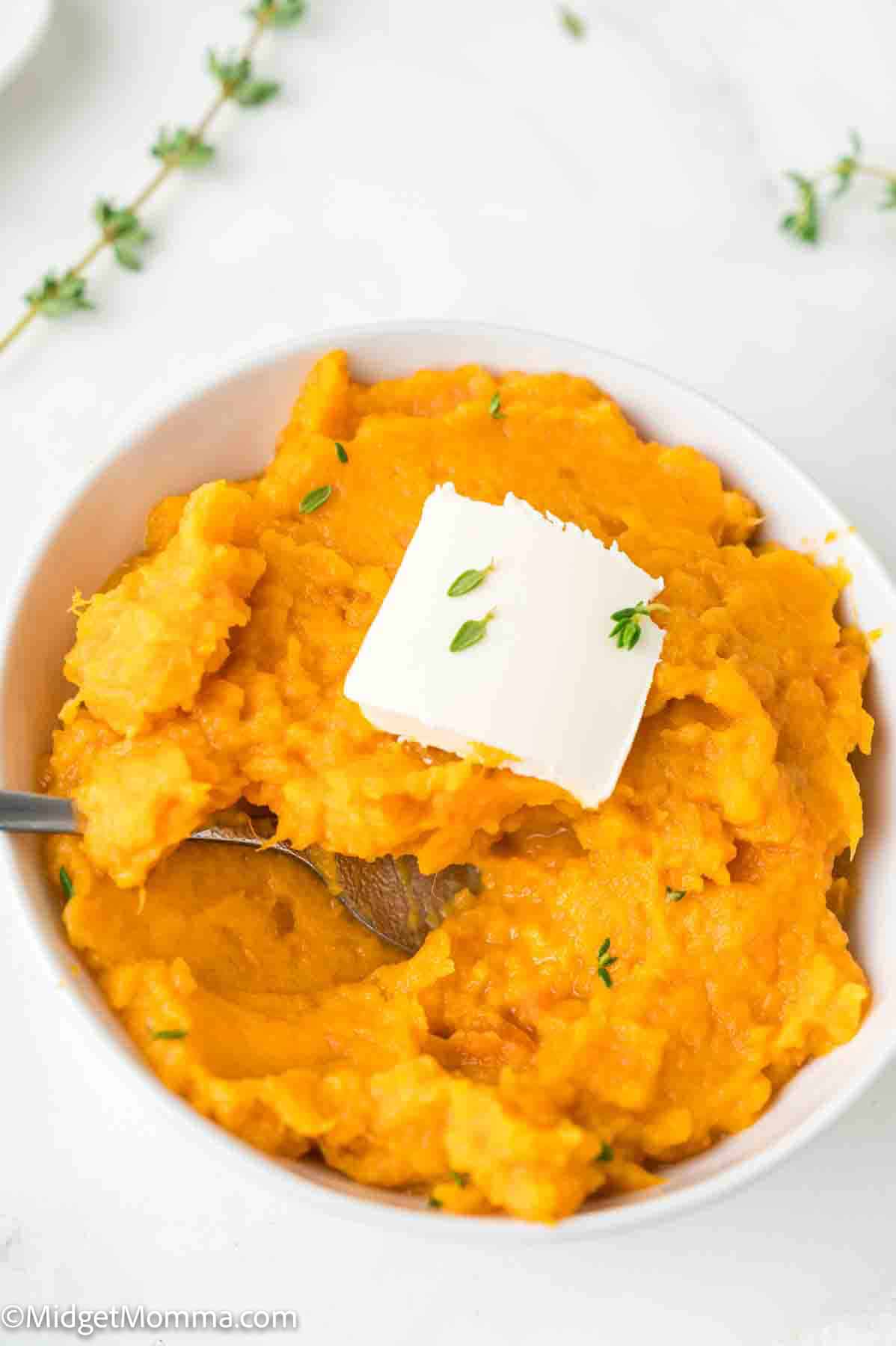 Bowl of mashed sweet potatoes with a pat of butter and thyme garnish on top. A spoon is partially submerged in the mash.