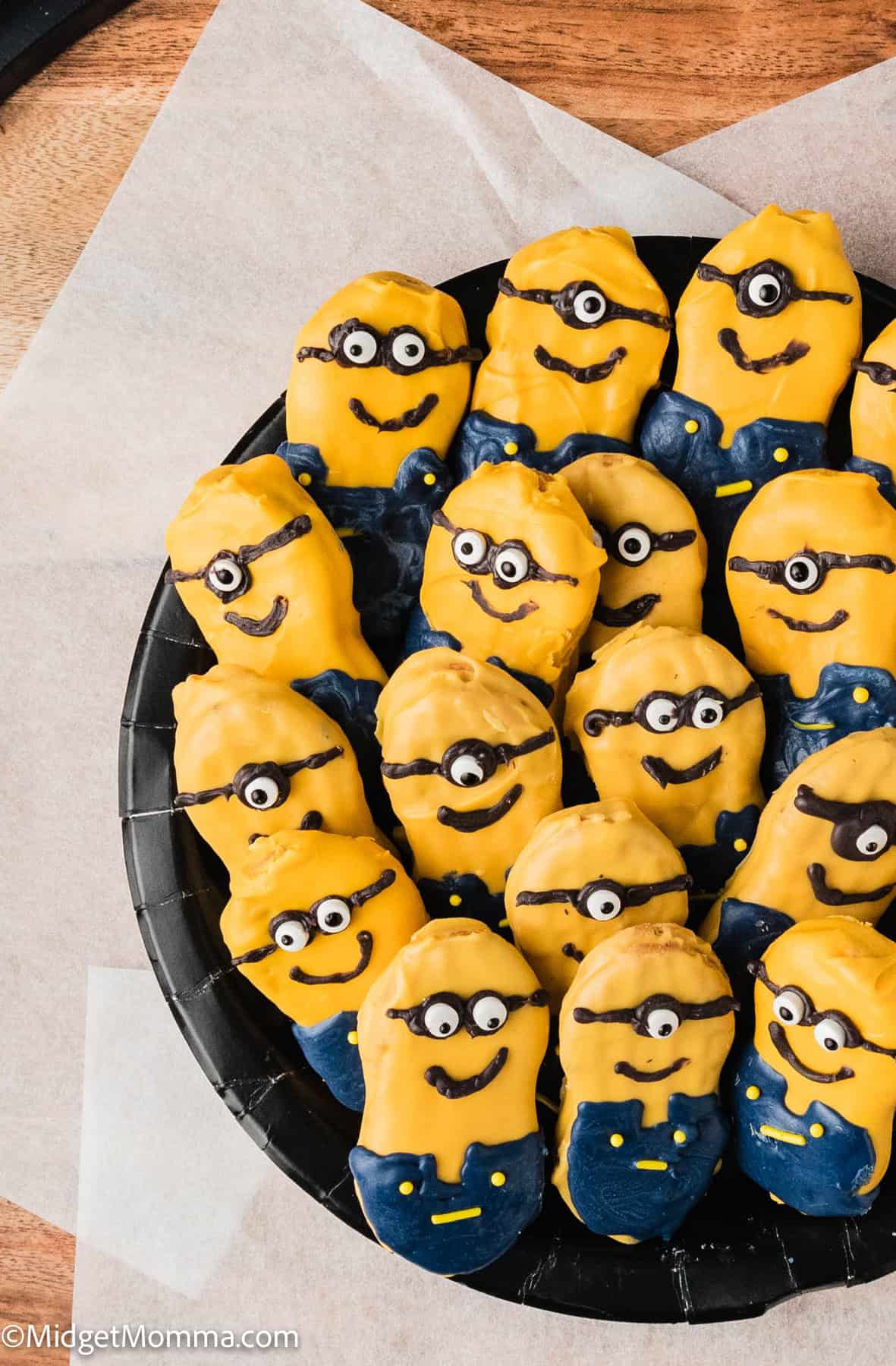 A plate of cookies shaped like cartoon minions, decorated with yellow and blue icing, arranged neatly on a black plate with a wood background.