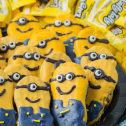 Cookies decorated as characters with yellow and blue icing, resembling minons, a popular animated character, displayed on a plate with yellow candy wrappers.