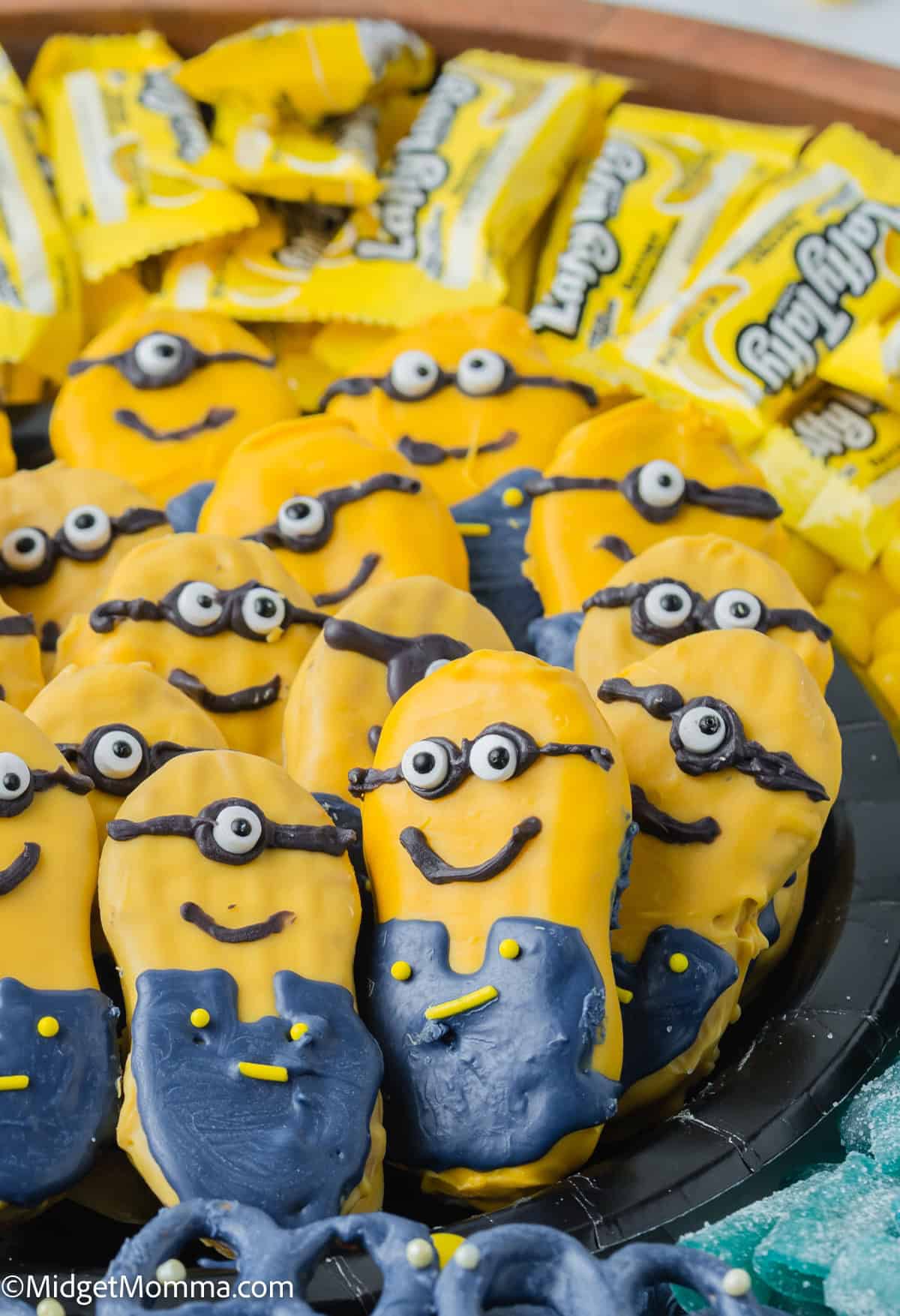 Cookies decorated as characters with yellow and blue icing, resembling minons, a popular animated character, displayed on a plate with yellow candy wrappers.