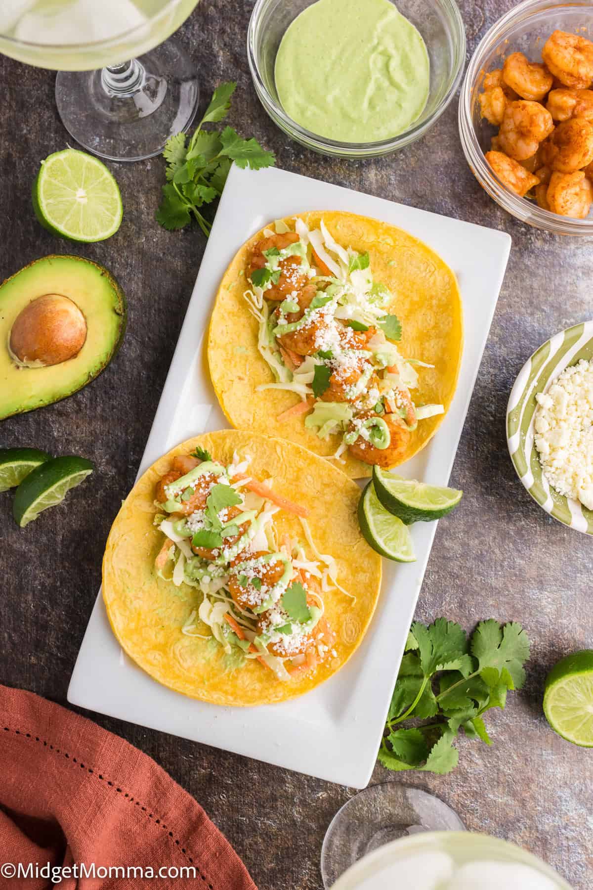 Two tacos on a white plate topped with shredded lettuce, cheese, and cilantro. Surrounding ingredients include avocado, lime, shrimp, and sauces.