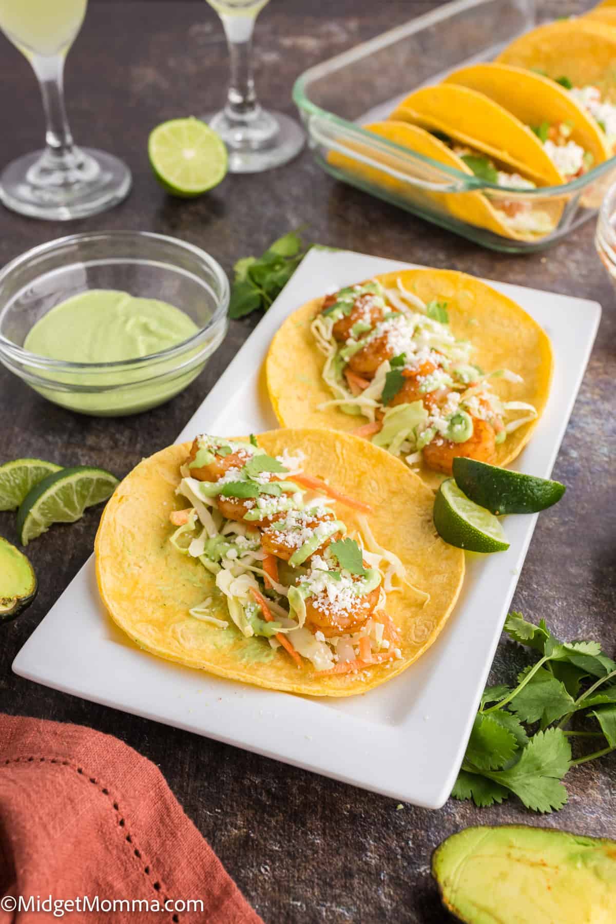 Two tacos on a rectangular white plate, topped with shredded lettuce, cheese, and sauce. Lime wedges are nearby. A glass dish with more tacos and a lime are in the background.