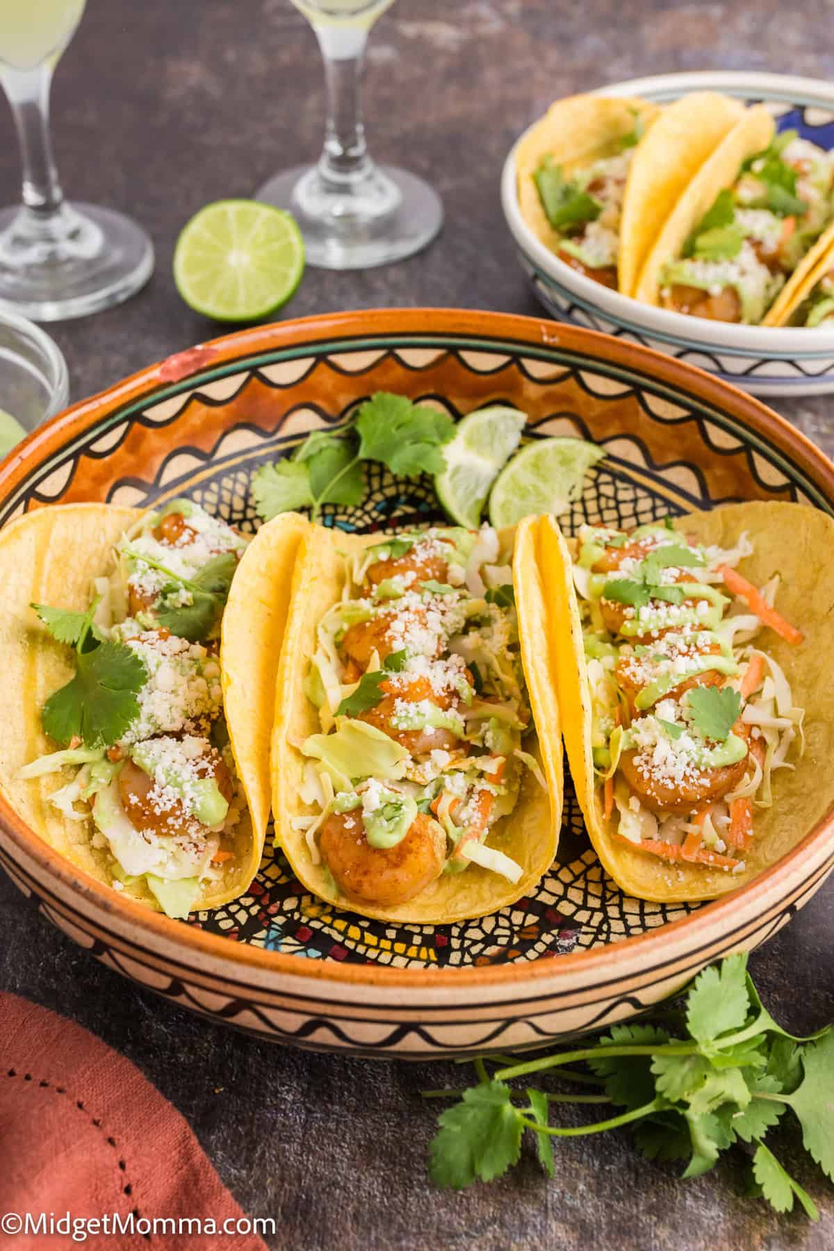 Three shrimp tacos topped with cabbage, cheese, and cilantro on a decorative plate, with a lime and more tacos in the background.