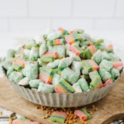 A metal bowl filled with green coated cereal pieces and colorful gummy candies, placed on a wooden board with gold candy sprinkles scattered around.