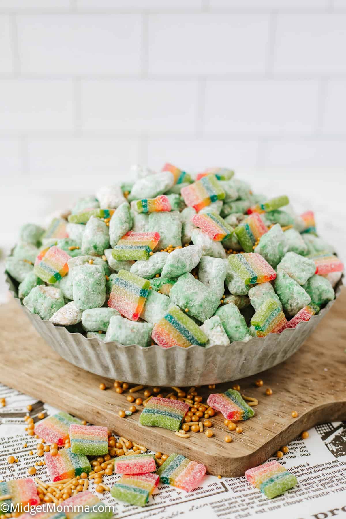 A metal bowl filled with green coated cereal pieces and colorful gummy candies, placed on a wooden board with gold candy sprinkles scattered around.