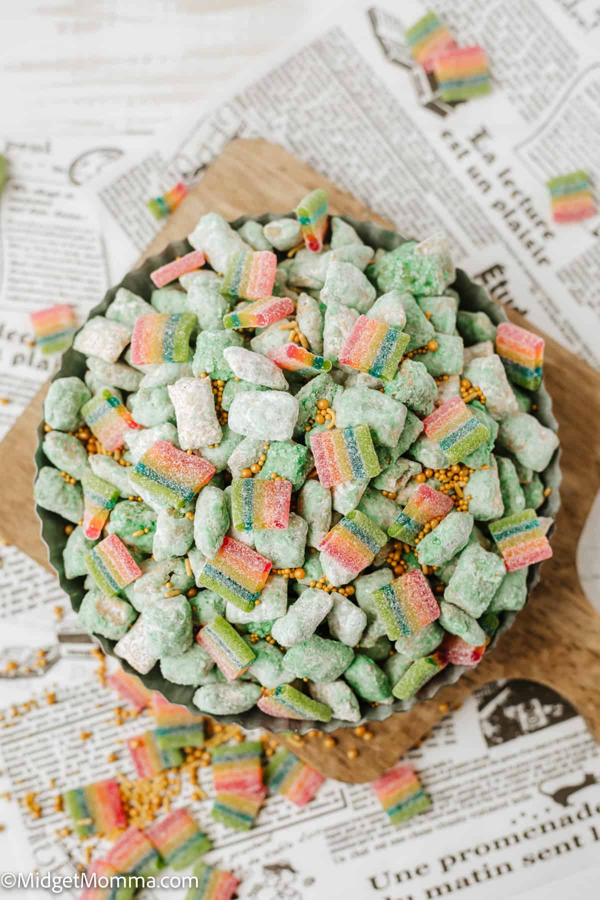 A bowl of green, powdered sugar-coated snacks with rainbow-striped gummy pieces and gold sprinkles, placed on a newspaper background.